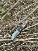 The Texan Knives 9" Damascus steel hunting knife with sheep horn/resin handle and leather sheath is displayed outdoors on dry twigs and grass.