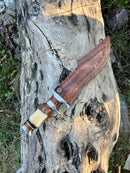 The Texan Knives Damascus Steel Sub-Hilt Fighter Bowie Knife 14” with leather sheath rests on a weathered log among grass and rocks—an ideal premium Christmas gift for outdoor enthusiasts.