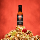 A bottle of Bravado Spice Co Garlic & Árbol Moruga Scorpion Hot Sauce, featuring the intense heat of Trinidad Moruga Scorpion peppers, rests on a stack of burritos topped with lime and a red pepper, all set against a vivid red backdrop—an ideal image to capture this ultra-hot Bravado Spice Co experience.