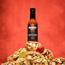 A bottle of Bravado Spice Garlic & Árbol Moruga Scorpion Hot Sauce, crafted with intense Trinidad Moruga Scorpion peppers and fiery árbol chiles, is balanced atop a pyramid of burritos stuffed with chicken and vegetables, garnished with lime wedges and a red chili pepper, set against a bold red background—the ultimate street taco companion.