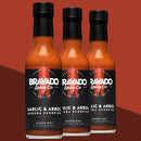 Three bottles of Bravado Spice Co Garlic & Árbol Moruga Scorpion Hot Sauce are arranged against a vivid red backdrop. Each bottle showcases a black label accented by white and red text, emphasizing the bold flavors and intense heat derived from Trinidad Moruga Scorpion peppers in this ultra-hot sauce by Bravado Spice Co.