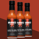 Three bottles of Bravado Spice Garlic & Árbol Moruga Scorpion Hot Sauce by Bravado Spice, featuring the fiery Trinidad Moruga Scorpion pepper, are displayed against a red background. The black labels and vibrant orange-red color of this ultra-hot sauce stand out—perfect for pairing with adventurous street tacos.