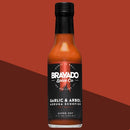 A bottle of Bravado Spice Co Garlic & Árbol Moruga Scorpion Hot Sauce, featuring fiery Trinidad Moruga Scorpion peppers and a sleek black label from Bravado Spice Co, sits against a bold red and dark red diagonal background—an ultra-hot sauce crafted for serious heat enthusiasts.