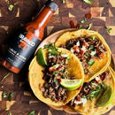 Three beef tacos topped with chopped onions, cilantro, and lime wedges sit on a wooden board beside a bottle of Bravado Spice Co Garlic & Árbol Moruga Scorpion Hot Sauce from Bravado Spice Co—an ultra-hot sauce made with fiery Trinidad Moruga Scorpion peppers.