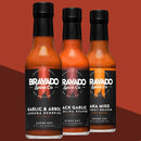 Three bottles of Bravado Spice hot sauce—featuring Garlic & Árbol Moruga Scorpion, Black Garlic Carolina Reaper, and Aka Miso Ghost-Reaper varieties—are displayed against a rich red and brown backdrop. This ultra-hot hot sauce collection from Bravado Spice is perfect for adventurous flavor seekers or those looking to elevate their next street taco experience.