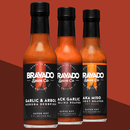 Three bottles of Bravado Spice hot sauce—featuring Garlic & Árbol Moruga Scorpion, Black Garlic Carolina Reaper, and Aka Miso Ghost-Reaper varieties—are displayed against a rich red and brown backdrop. This ultra-hot hot sauce collection from Bravado Spice is perfect for adventurous flavor seekers or those looking to elevate their next street taco experience.