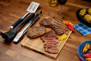 A wooden board with grilled steaks, peppers, and Hasselback potatoes sits next to the Napoleon Grills 3-Piece Starter Tool Set Essential Series (GATL002), a glass of red wine, and oil—perfect for starting your grilling journey.