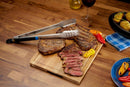 Grilled steak on a wooden cutting board with slices arranged, topped with Napoleon Grills' Essential Series Grill Tongs (GATL004). Roasted peppers and a glass of red wine are nearby, along with a plate of food and potatoes in the background.