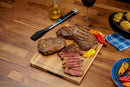 Grilled steaks and bell peppers rest on a wooden board. The scene includes the Napoleon Grills Silicone Basting Brush Essential Series (GATL005), a jug of oil, red wine, and plates of roasted potatoes and peppers.