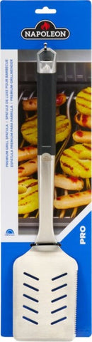 The Napoleon Grills Premium Grill Spatula PRO Series (GATL007) features a stainless steel slotted blade and a black handle, shown in packaging with grilled hot dogs and vegetables in the background—ideal for grilling enthusiasts.