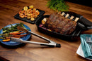 A roasted rack of ribs in a black pan, grilled shrimp and veggies on a black plate, and broccolini with carrots on blue plate. Napoleon Grills Premium Grill Tongs PRO Series (GATL008) with QuickClick locking rest nearby on the wooden table.