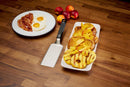A plate with bacon and fried eggs sits next to a platter of French toast topped with grilled peaches. The Napoleon Griddle Spatula for Griddles (GATL014) by Napoleon Grills and fresh peaches are also on the wooden table.