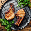 Two grilled pork chops with char marks rest on a dark plate, garnished with fresh parsley and SALTOPIA Infused Sea Salts' Applewood Smoked Salt: GREAT OUTDOORS, atop a rustic wooden table.
