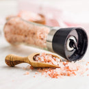 A wooden scoop and a grinder, both filled with SALTOPIA Infused Sea Salts' Saltopia Pink Himalayan Coarse Salt 3lbs, rest on a surface sprinkled with scattered pink salt crystals.