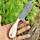 A hand holds the Texan Knives Gareth Bull Shamwari Inspired 8" Handmade Damascus Knife with a camel bone handle and leather sheath, set against green leaves and a tree trunk.
