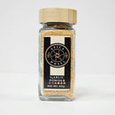 A glass jar of Spice Wheel Garlic Powder by Spice Wheel, crafted from premium dehydrated garlic and topped with a wooden lid, is labeled “Garlic Powder Net Wt. 65g” against a plain white background. This essential pantry staple imparts rich, savory flavor to any dish.