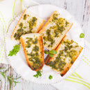 Four slices of garlic bread topped with SALTOPIA Infused Sea Salts' PRESTO PESTO Basil, Garlic & Lemon Salt are arranged on a white plate, set on a rustic white wooden surface with a white and green striped cloth beside it.