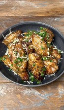 A black plate with glazed chicken wings, topped with Terrapin Ridge Farms Truffle Hot Sauce, shredded cheese, and fresh parsley, sits on a rustic wooden table.