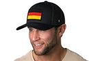 A man in a light shirt smiles against a white background, wearing the Haka Hat German Flag Trucker Hat—Black Germany Snapback, featuring an embroidered Germany flag patch on the front.