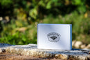 A white gift box bearing the Brushfire Farms logo rests on a stone surface outdoors, showcasing their El Jefe Bundle, with lush green foliage softly blurred in the background.