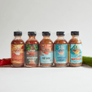 Five bottles from the Culture Hot Sauce Gift Sampler pack by Culture Hot Sauce, each featuring a vibrant label, are arranged in a row for an attention-grabbing display. The sampler showcases bestselling flavors—Blindsider, Hungarian Secret, The Don, Tahitian Dream, and Bar Sauce. Fresh red and green peppers flank the pack, emphasizing this colorful set that makes a perfect gift for any spice enthusiast.