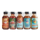 Five bottles from the Culture Hot Sauce Gift Sampler pack by Culture Hot Sauce, each featuring a vibrant label, are arranged in a row for an attention-grabbing display. The sampler showcases bestselling flavors—Blindsider, Hungarian Secret, The Don, Tahitian Dream, and Bar Sauce. Fresh red and green peppers flank the pack, emphasizing this colorful set that makes a perfect gift for any spice enthusiast.