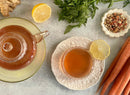 A cup and teapot of World Spice Ginger Sunrise herbal tea with a lemon slice, surrounded by fresh carrots, a lemon half, carrot greens, ginger, and a dish of dried tea herbs from World Spice on a light surface evoke the vibrant flavors of lemon and ginger.