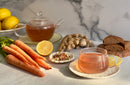 A glass teapot and cup filled with World Spice Ginger Sunrise herbal tea—by World Spice—are surrounded by fresh carrots, ginger root, a halved lemon, dried herbs, lemons in a bowl, and slices of brown bread on a marble countertop.
