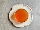 A clear glass cup filled with vibrant World Spice Ginger Sunrise tea sits on a white, beaded-edge plate atop a gray textured surface.