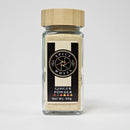 A glass jar with a wooden lid, branded Spice Wheel and labeled "Spice Wheel Ginger Powder," Net Wt. 50g, holds light beige powdered ginger with a zesty aroma and is displayed on a white surface against a plain white background.