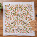 The Gingiber Apple Blossom Tea Towel by Gingiber features a symmetrical bird, pink flower, and green leaf design. Shown on wood beside tan gardening gloves, a small plant, and part of a garden tool. Made from square flour sack cotton.