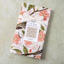 A neatly folded Gingiber Apple Blossom Tea Towel, featuring a floral and bird pattern, is tied with a white ribbon on a light textured surface. The label displays the brand Gingiber and a sample of the design.