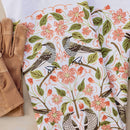 A pair of brown gardening gloves beside the Gingiber Apple Blossom Tea Towel, featuring pink flowers, green leaves, and black-and-white birds on a white background.