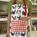 Wearing a green shirt and the Gingiber Chicken Apron—an adjustable black and white chicken design with red checkered pockets from Gingiber—a person stands by fabric shelves, holding a beige tote bag.