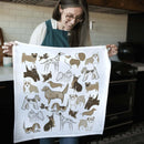 A woman in glasses and an apron holds up the Gingiber Dog Tea Towel by Gingiber, featuring cartoon illustrations of various dog breeds in brown, tan, and black on white flour sack cotton. She stands in a kitchen.