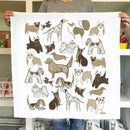 A person holds up the Gingiber Dog Tea Towel by Gingiber, a white flour sack cotton towel featuring playful, cartoon dog breed illustrations in browns, tans, and blacks. Colorful items are visible on shelves in the background.