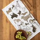 The Gingiber Dog Tea Towel by Gingiber, featuring illustrations of various dog breeds in neutral colors, rests on a wooden surface next to a brown bowl of green grapes and two wooden utensils, adding charm to your kitchen.