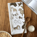 The Gingiber Dog Tea Towel by Gingiber, made from flour sack cotton and featuring illustrated dogs in brown, tan, and black, is perfect for kitchen cleanup. Shown on a wooden table with a spoon, measuring cup of sugar, open book, and bowl of oats.