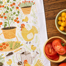 The Gingiber Garden Chicken Tea Towel by Gingiber is a white flour sack cotton towel with a colorful garden print featuring chickens, tomato plants, potted flowers, and a watering can, shown beside bowls of yellow and red tomatoes.