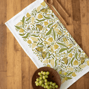 The Gingiber Golden Garden Tea Towel by Gingiber, crafted from soft flour sack cotton, features a charming yellow and green floral and butterfly design. It’s displayed on wood beside wooden utensils and a bowl of green grapes.