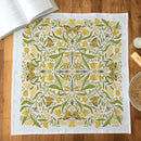 The Gingiber Golden Garden Tea Towel by Gingiber features a symmetrical yellow and green botanical design with butterflies, flowers, and leaves. Shown flat on wood, it's surrounded by a book, bowl, and small items.