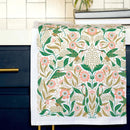 The Gingiber Hummingbird Tea Towel by Gingiber features green birds, leaves, and pink flowers on soft flour sack cotton, and is shown draped over a dark blue cabinet with a gold handle beside a light counter and tiled backsplash.