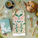 A flat lay of a tea setting with a copper teapot, two cups, wooden spoons, tea bags, corks, the Gingiber Hummingbird Tea Towel by Gingiber featuring birds and florals, and a Little Women book on a green textured background.