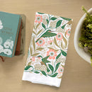 A folded Gingiber Hummingbird Tea Towel by Gingiber, with a vibrant floral and hummingbird design, rests on a light wooden table beside a stack of books and a white bowl filled with green foliage.