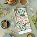 A flat lay shows a tea setting with cups, tea bags, flowers, and the Gingiber Hummingbird Tea Towel by Gingiber—a decorative cotton towel featuring green birds and pink flowers—on a green surface, with a wooden spoon resting on the towel.
