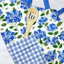 The Gingiber Hydrangea Apron by Gingiber features a blue and white cotton design with hydrangea and bee pattern, blue straps, a blue gingham towel draped over it, and a wooden slotted spoon in the pocket.