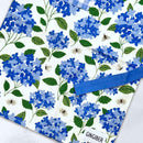 The Gingiber Hydrangea Apron by Gingiber is a cotton kitchen apron with blue hydrangea flowers, green leaves, and bees on white. It features a blue ribbon, a GINGIBER tag, and is adjustable for the perfect cooking fit.