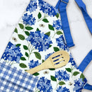 The Gingiber Hydrangea Apron by Gingiber features blue and white cotton with hydrangea and bee designs, plus a blue plaid pocket. It’s shown with a wooden slotted spoon on a white surface.