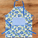 The Gingiber Hydrangea Apron by Gingiber showcases blue hydrangea patterns and a blue gingham pocket. Made of cotton, it features adjustable blue neck and waist ties for comfort and style, displayed on a wooden surface.