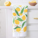 The Gingiber Lemon Grove Tea Towel, made of flour sack cotton and featuring yellow lemons and green leaves with "GINGIBER" at the bottom, hangs over a white drawer near a halved lemon and a bowl of lemons.